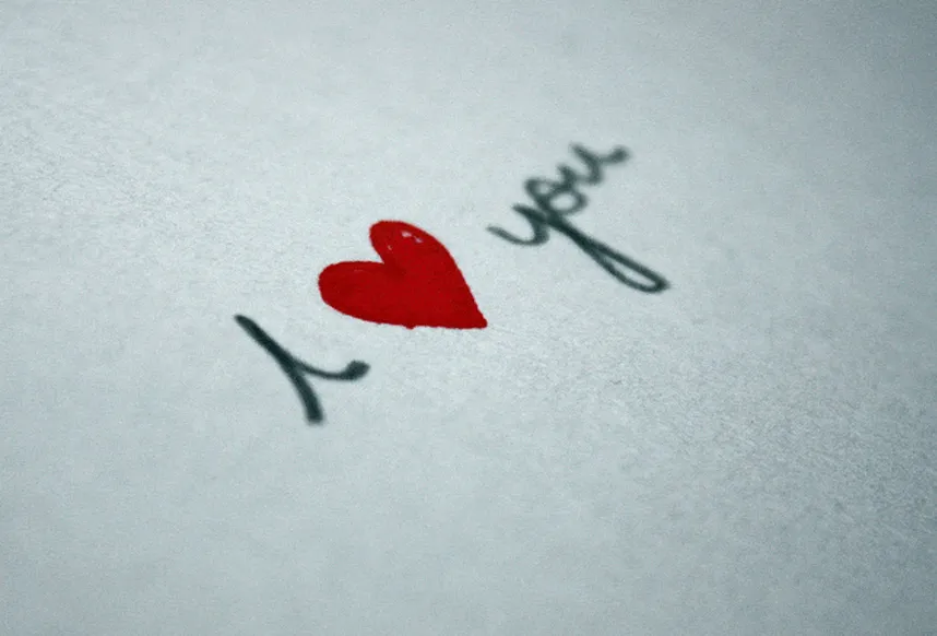 Текст на белом листе бумаги. Красное сердце. В переводе: Я люблю тебя!